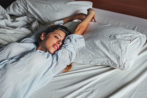 Blonde woman in a striped button down stretching in bed, sunlight beaming in. Blonde woman in a striped button down stretching in bed, sunlight beaming in.